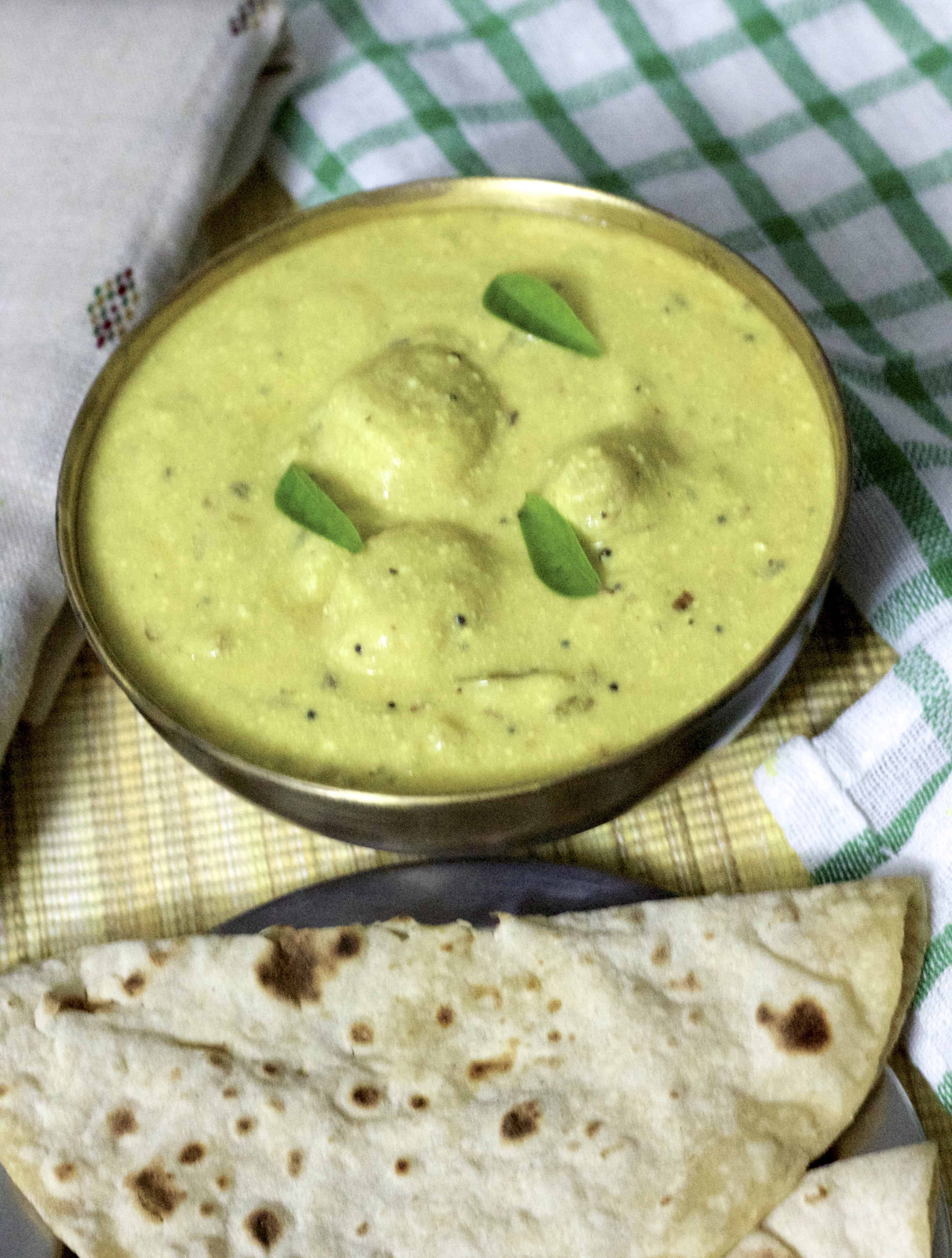 Brass bowl with yellow arbi ki kadhi/ colocasia yogurt gravy garnished with curry leaves, a plate of chappaties, a white and green checked napkin, all on a cane mat and a beige hand embroidered shawl on the left