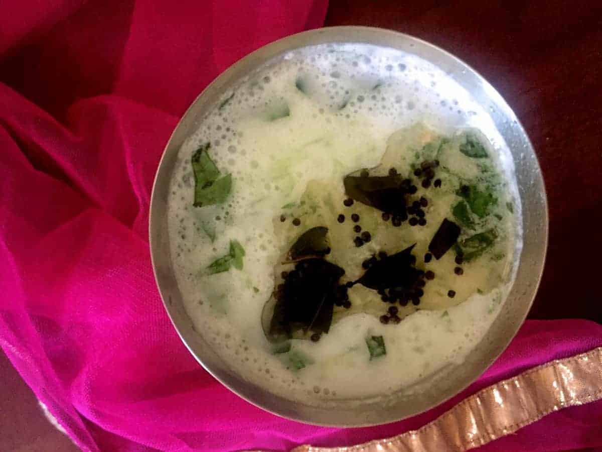 Neer Mor a mildly spiced and herbed buttermilk summer cooler garnished with curry leaves. Resting on a gauzy pink fabric. Served in a tall glass with cucumber garnish and tempered with mustard seeds and curry leaves