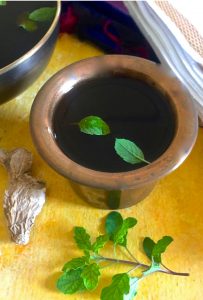 Brass tumbler with cooling dark jaggery drink Panakam with tulsi leaves as garnish, on a yellow background