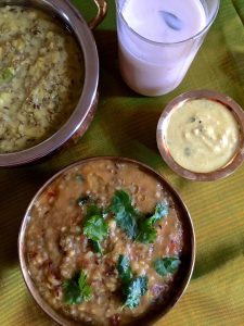 A bowl of greenish yellow cooked millets and lentils with a glass of buttermilk and the edge of an orange book at one corner