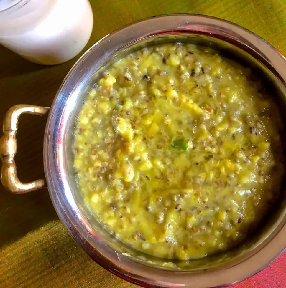 A brass bowl with an handle to the left filled with greenish brown Bajra Khichdi of Pearl millets and a glass of buttermilk by the side