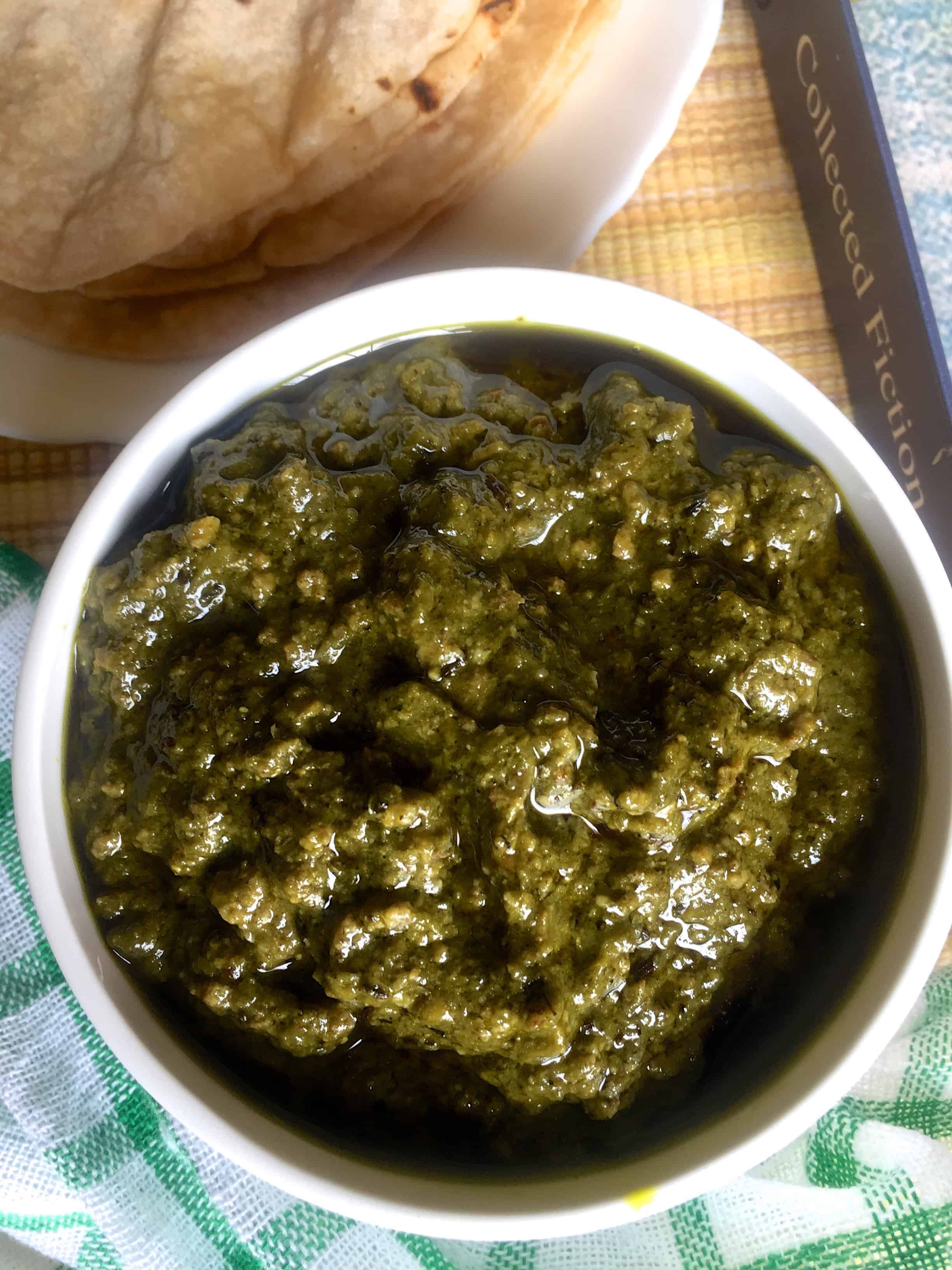 A white bowl of greenish sept vadi curry with a plate of chaptatis and green checked napkin alongside