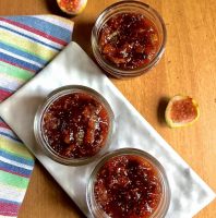 3 little jars of reddish brown Homemade fig jam with mint and no pectin on a white rectangular marble slab, with fig halves on one side and a napkin with blue, yellow, red and green stripes on the left