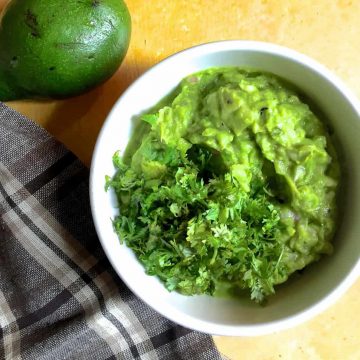 A white bowl with fresh homemade guacamole garnished with cilantro leaves. A checked grey and brown napkin to the left and part of an avocado seen on the top left corner