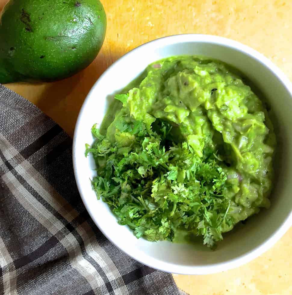 A white bowl with fresh homemade guacamole garnished with cilantro leaves. A checked grey and brown napkin to the left and part of an avocado seen on the top left corner