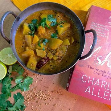 Panch Phoron Kaddu Sabzi, chunks of spiced pumpkin curry in an iron pan with a red book on Emperor Ashoka on one side and yellow mustard and cilantro scattered below
