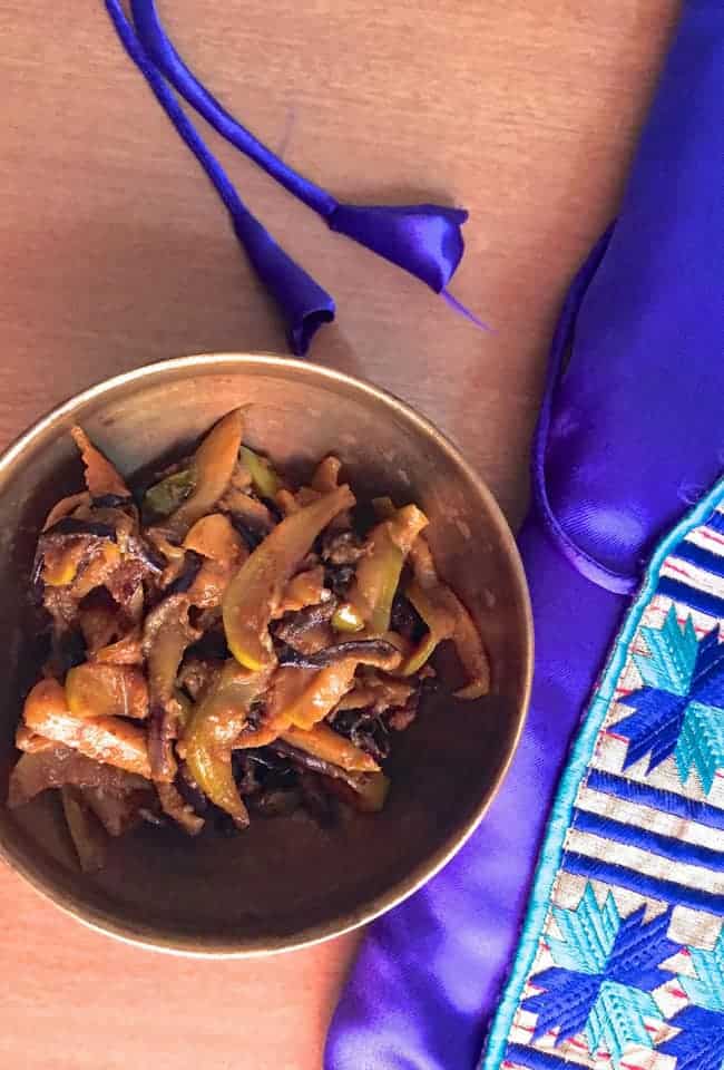 Kashmiri Green Apple Baingan Curry in a brass bowl on a pale brown background and a silk embroidered jacket alongside in purple and blue