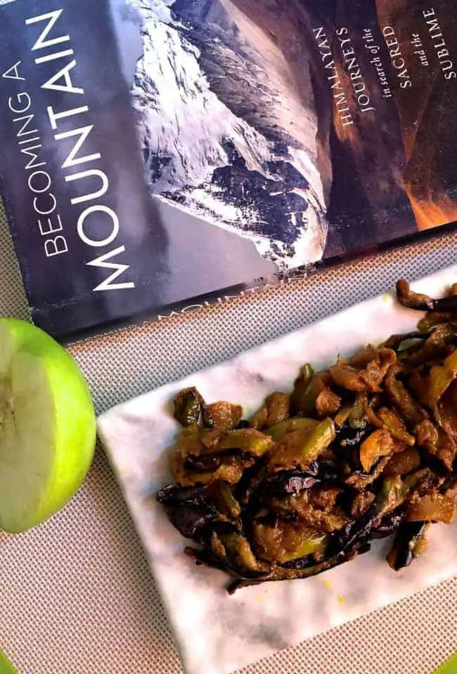 Fried eggplant with green apples cooked in spices, on a white marble slab, a slice of green apple on the left and a book on the Himalaya mountains above it