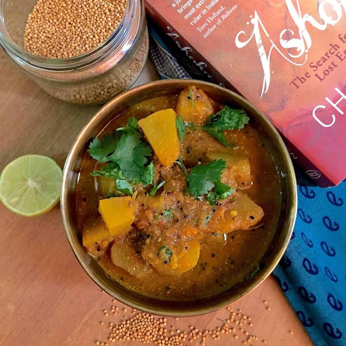 Panch Phoron Kaddu Sabzi, chunks of spiced pumpkin curry in an iron pan with a red book on Emperor Ashoka on one side and yellow mustard and cilantro scattered below