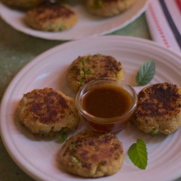 White plates with little brown aloo tikkis or potato patties, with a little cup of red tamarind chutney and mint leaves