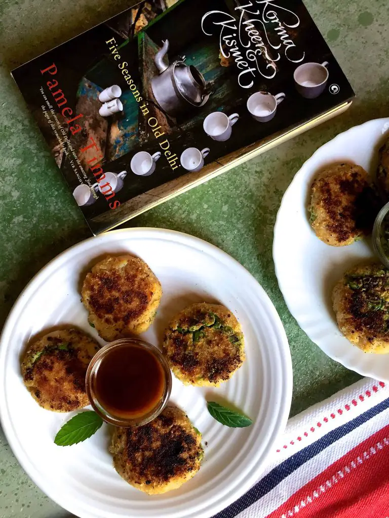 4 aloo tikki or potato patties on a white plate with mint leaves between and a book Korma Kheer Kismet alongside