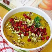 Curried Roasted Pumpkin Soup with apples and Indian spices including turmeric, in a white bowl and a garnish of ruby red pomegranate arils and crushed pista nuts