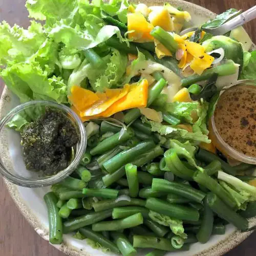 A bowl of beans, greens and green and yellow zucchini ribbons with green pesto and orange citrus dressing in tiny cups on either side of the bowl to make up Pesto Beans Zucchini Salad with Citrusy dressing
