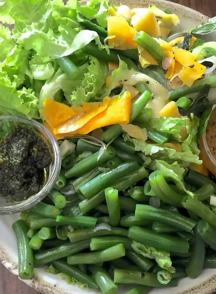 A bowl of beans, greens and green and yellow zucchini ribbons with green pesto and orange citrus dressing in tiny cups on either side of the bowl to make up Pesto Beans Zucchini Salad with Citrusy dressing