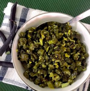 Sliced sauteed Purple Beans Poriyal garnished with grated coconut in a white bowl with a fork stuck in at one side. Resting on a white an black striped napkin all on a dark green background