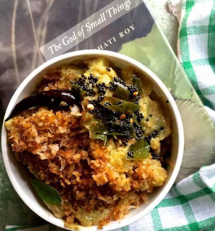 A bowl of Raw Banana Varutha Erissery, a curry of green plantain cooked in turmeric and roasted coconut paste, resting on a green and gray book and a white and green checked napkin on the right