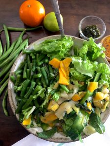 A bowl of Pesto Beans Zucchini Salad with fresh green beans, ribbons of zucchini, salad greens and bowls of green pesto and an orange citrus dressing to the right, an orange and a lemon behind the salad bowl and green beans on the left