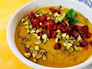 Yellow curried roasted pumpkin soup in a white edged bowl with a garnish of ruby red pomegranate arils and greenish yellow pista nuts with sprig of parsley alongside