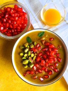 A bowl of pumpkin soup of roasted pumpkin curried with Indian spices, garnished with pomegranate and pista nuts and with a cup of orange juice on a white napkin and a bowl of pomegranates alongside
