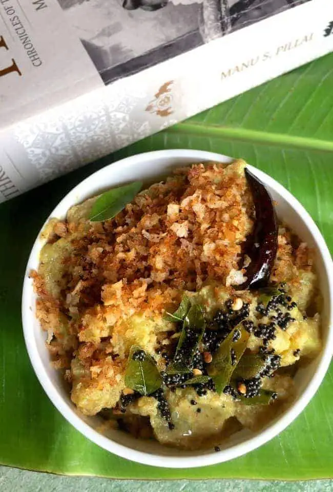 Raw Banana Varutha Erissery in. white bowl on a green plantain leaf, with a book on Kerala History in the background