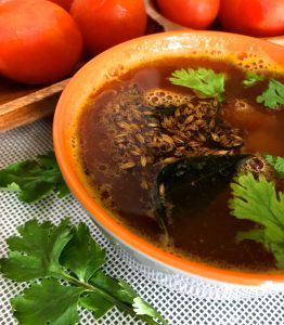 Tomato Rasam in an orange bowl, garnished with coriander leaves and tempered with mustard seeds and cumin, with a row of tomatoes behind