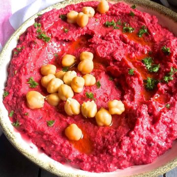 A large bowl of roasted beetroot hummus with the pink hummus garnished with chickpeas and bits of parsley and with olive oil