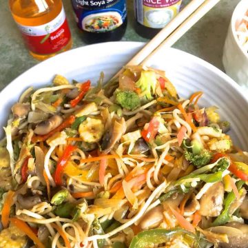 Stir fried vegetable noodles in a bowl with a pair of chopsticks and a glimpse of bottles of sesame oil, rice vinegar and soy sauce for the seasoning