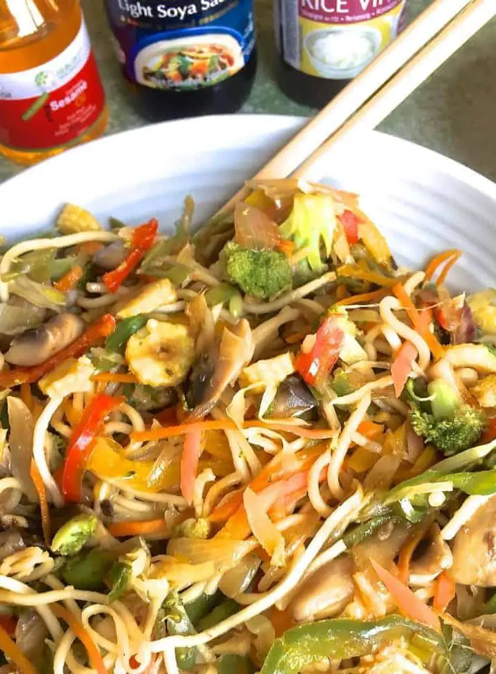 Stir fried vegetable noodles in a bowl with a pair of chopsticks and a glimpse of bottles of sesame oil, rice vinegar and soy sauce for the seasoning