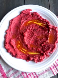 Pink and delicious looking roasted beetroot hummus spread on a white plate with olive oil shining on the hummus, a striped pink and white napkin alongside