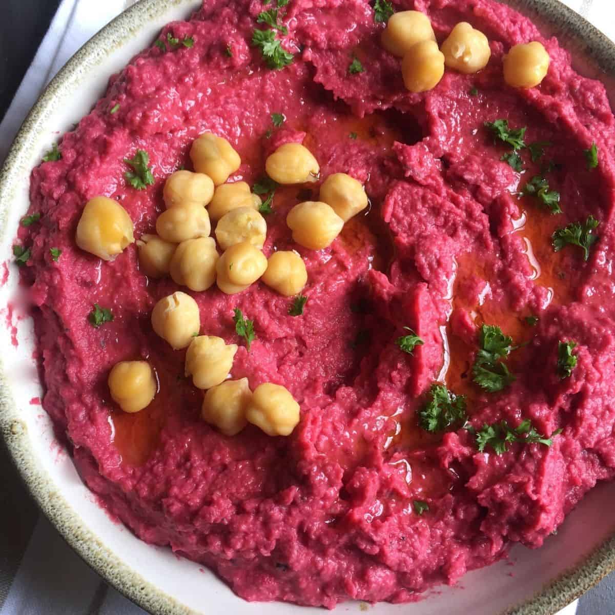 Bright bowl of roasted beetroot humus garnished with cooked chickpeas and olive oil, and with a basket of pita bread by the side