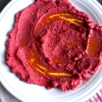 Pink and delicious looking roasted beetroot hummus spread on a white plate with olive oil shining on the hummus, a striped pink and white napkin alongside