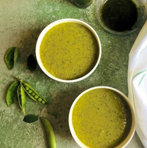Warming Pea Mint Basil Soup in two white bowls on a green background with split pea pods, mint and basil leaves by the side and a white napkin and a bowl of basil oil seen alongside