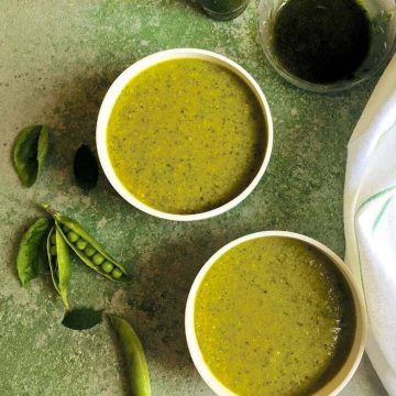 Warming Pea Mint Basil Soup in two white bowls on a green background with split pea pods, mint and basil leaves by the side and a white napkin and a bowl of basil oil seen alongside