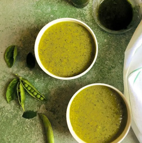 970×978 Pea Mint Basil Soup Warming Pea Mint Basil Soup in two white bowls on a green background with split pea pods, mint and basil leaves by the side and a white napkin and a bowl of basil oil seen alongside