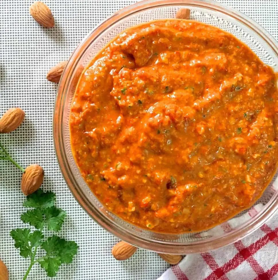 Spanish Romesco Sauce from charred red peppers almonds and tomatoes in a bowl resting on a red striped napkin to one side and with almonds and parsley scattered on the left