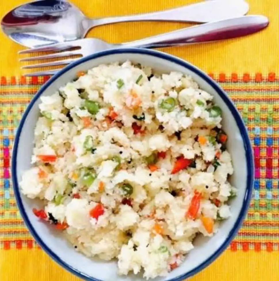 Hot tasty breakfast Vegetable rava Upma with semolina flour. A bowl of Upma on a yellow patterned runner with a steel fork and spoon above