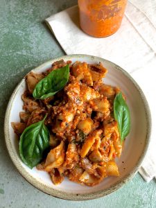 A bowl of shell pasta in an orangey red Romesco Sauce, with large basil leaves as garnish and a pale linen napkin on the side with a jar of the sauce