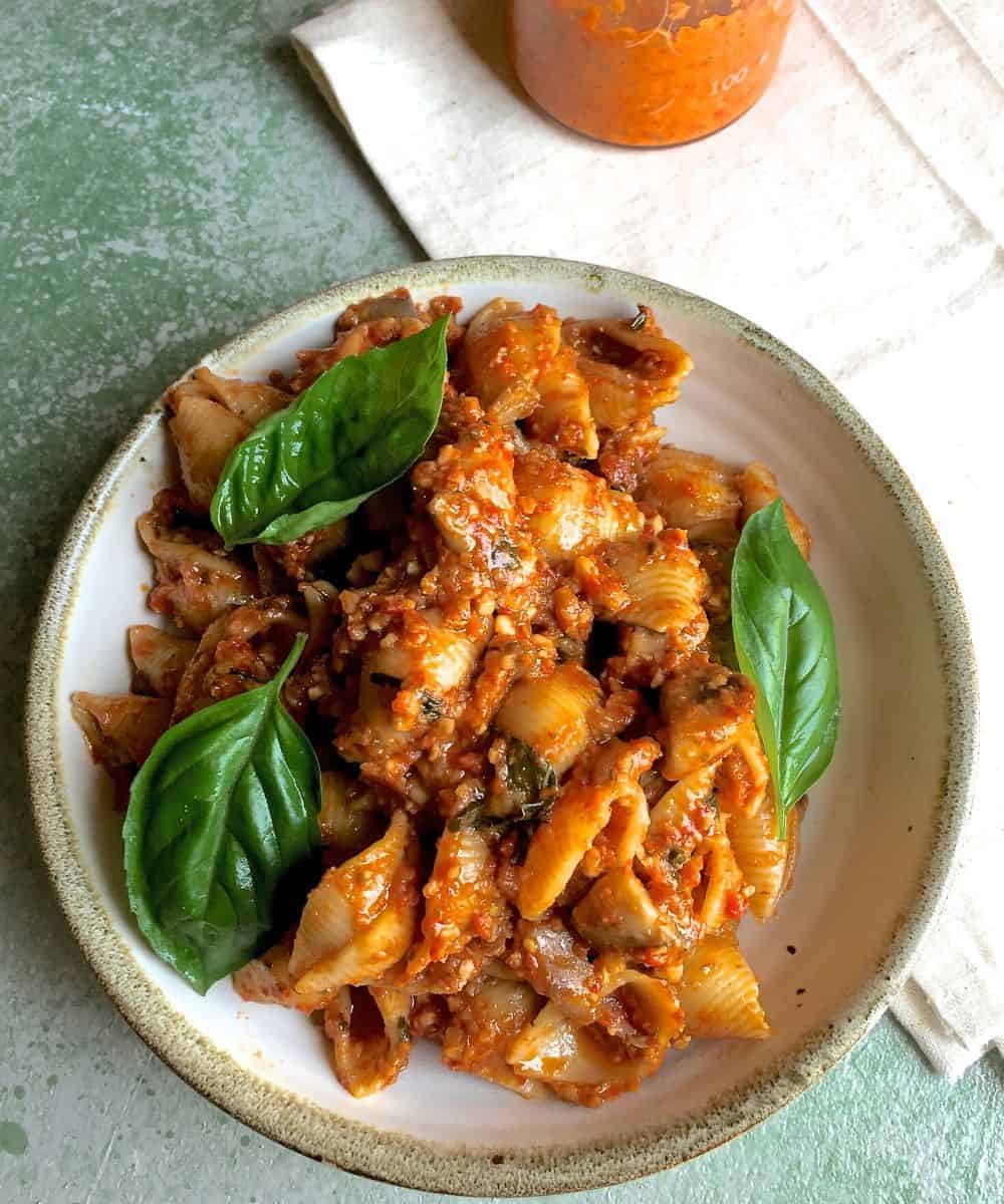 Beige edged plate filled with shell pasta in a an orangey pasta sauce with fresh basil leaves as garnish. A jar of sauce in the background on a white napkin, all of this on a green background