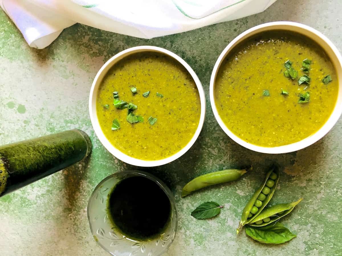 Two bowls of green Pea Basil and Mint soup with a bowl of basil oil and split green pea pods in the foreground, all on a green background and with a white napkin at the background