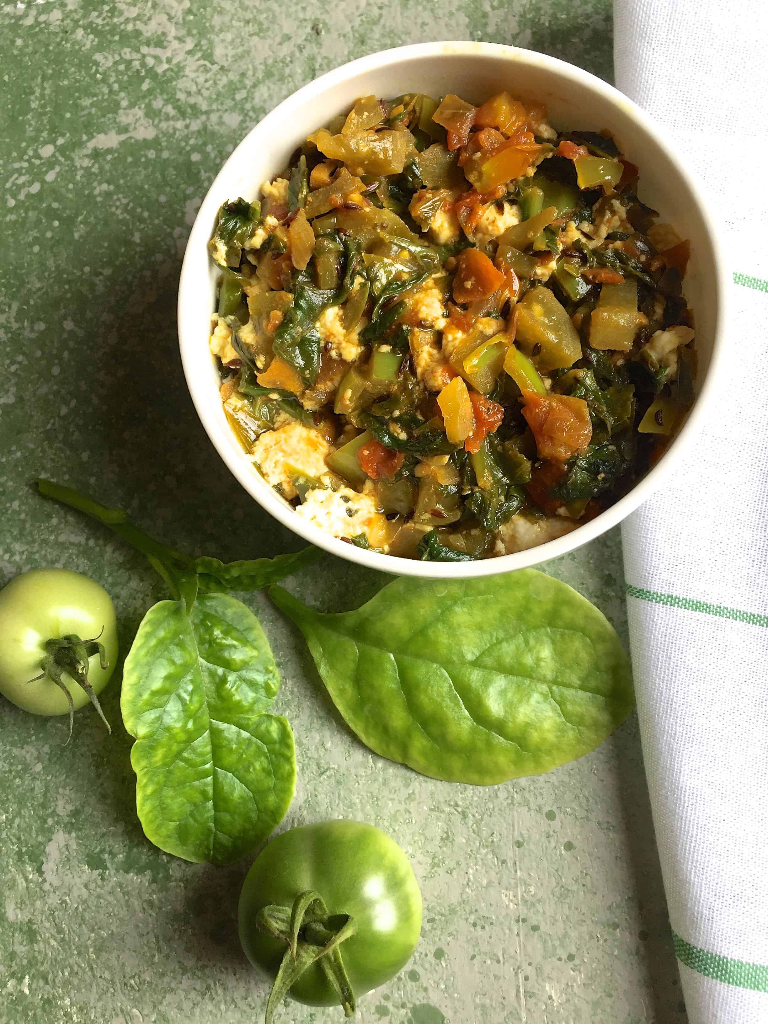 Radish Greens Spinach Curry in a white bowl with green tomatoes and Malabar spinach leaves on a green background and a white napkin striped with green on the side