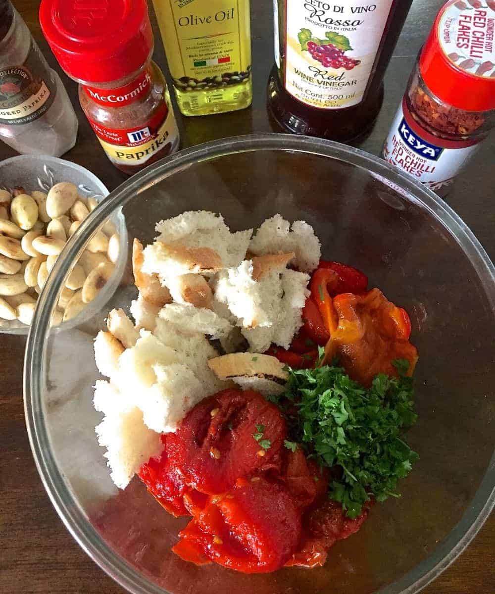 Ingredients for Spanish Romesco Sauce: Glass bowl with bread, roasted red peppers, parsley and tomatoes, with blanched peeled almonds in another bowl and other ingredients in bottles in the background