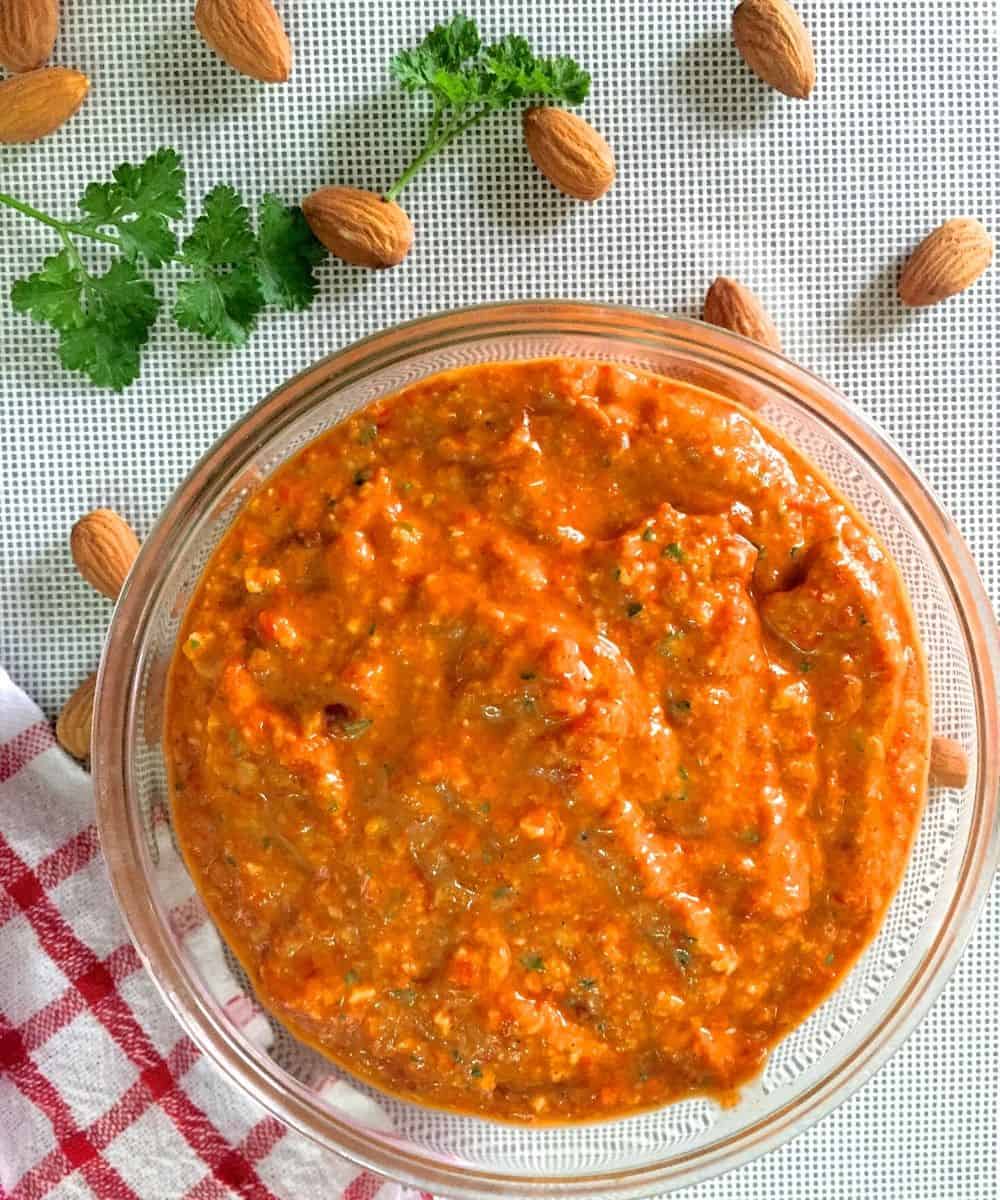 Orange Red thick Spanish Romesco sauce in a glass bowl with almonds and parsely scattered in the background. A red and white checked napkin lies to the left