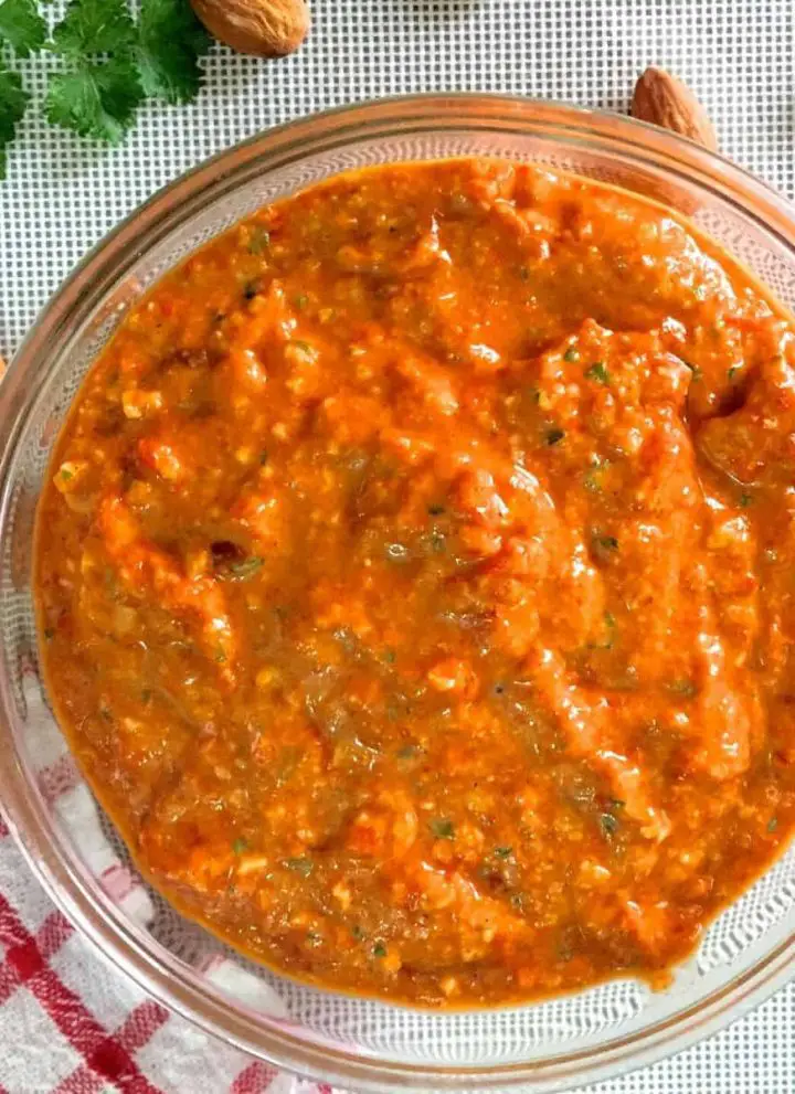 Glass bowl filled with bright orange thick Spanish Romesco Sauce with flecks of green from parsley. Almonds and parsley scattered in the background and a red and white checked cloth to the left