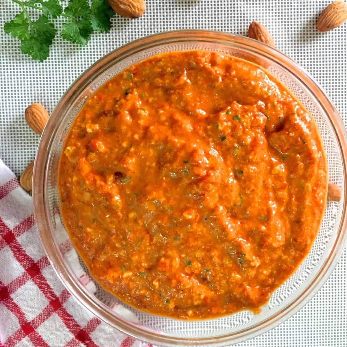 Glass bowl filled with bright orange thick Spanish Romesco Sauce with flecks of green from parsley. Almonds and parsley scattered in the background and a red and white checked cloth to the left