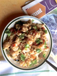 Vegetable Upma for breakfast with the days newspaper all handy. A white plate rimmed with green holds the Upma dotted with the red of tomatoes and the green of capsicum
