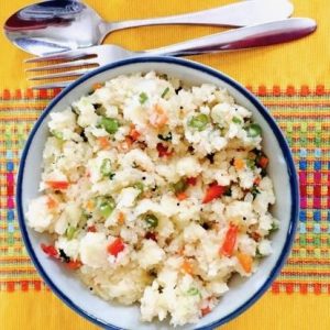 Vegetables cooked with sooji or semolina for a breakfast treat, in a bowl edged with blue on a bright yellow table cloth and a steel fork and spoon on top