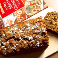 Brown coloured Eggless Christmas Cake with icing sugar strewn on top. A slice of cake lying on a wooden board, to the right and a red white and green book on Christmas cookies in the background