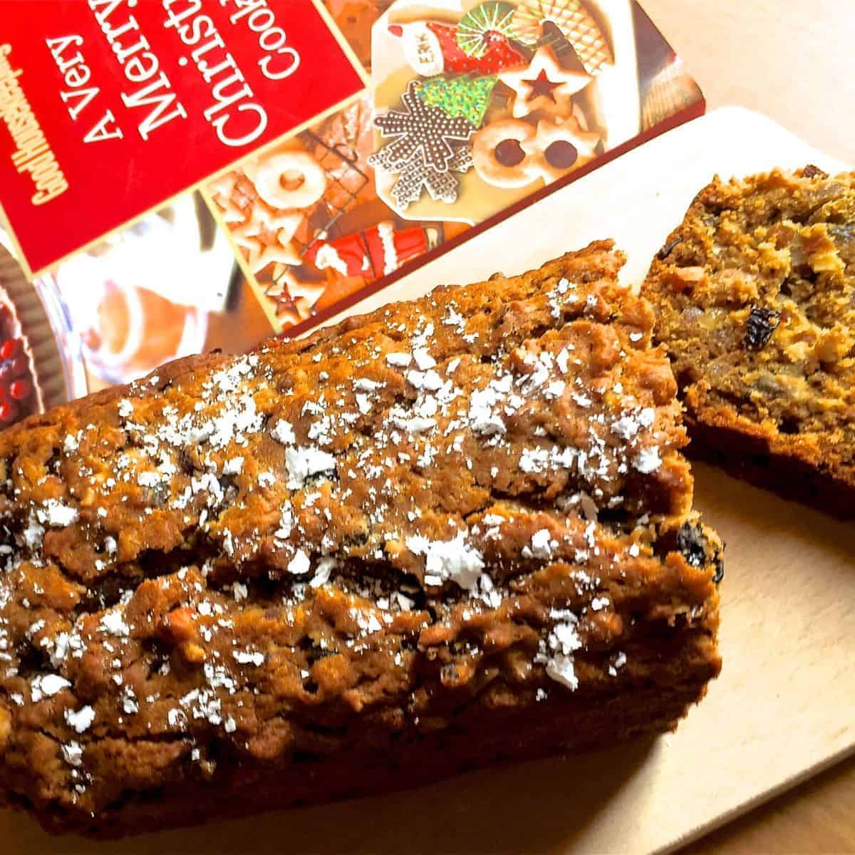 Dark Brown coloured Eggless Christmas Cake with icing sugar strewn on top. A slice of cake lying on a wooden board, to the right and a red white and green book on Christmas cookies in the background