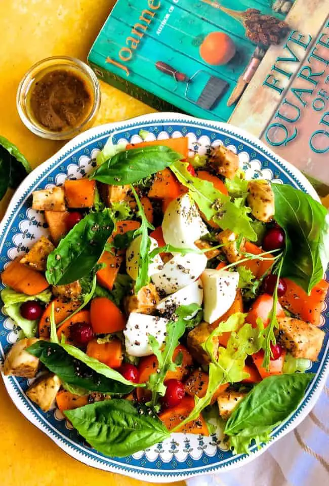 Orange green and white persimmon, mozzarella and basil make up this persimmon mozzarella salad shown on a blue edged plated with a green and orange book on one side and a bowl of mustard balsamic dressing on the other