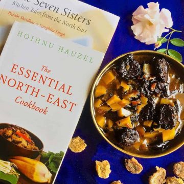 A brass bowl with aloo badi, a Manipuri dish of potatoes and little sundried nuggets of black gram lentil paste, on a purple background and book on the North East Indian cuisine alongside